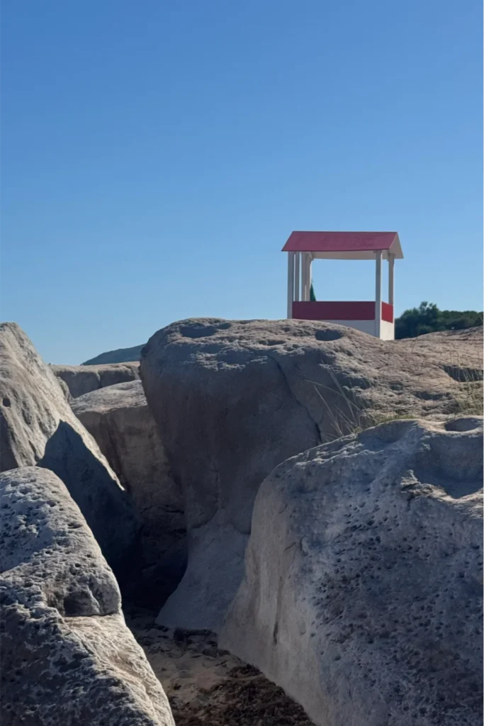 Casetta in legno sulla roccia Piccola struttura in legno bianca e rossa, simile a un gazebo da spiaggia, posta sopra una formazione di rocce granitiche chiare, contro un cielo azzurro senza nuvole.