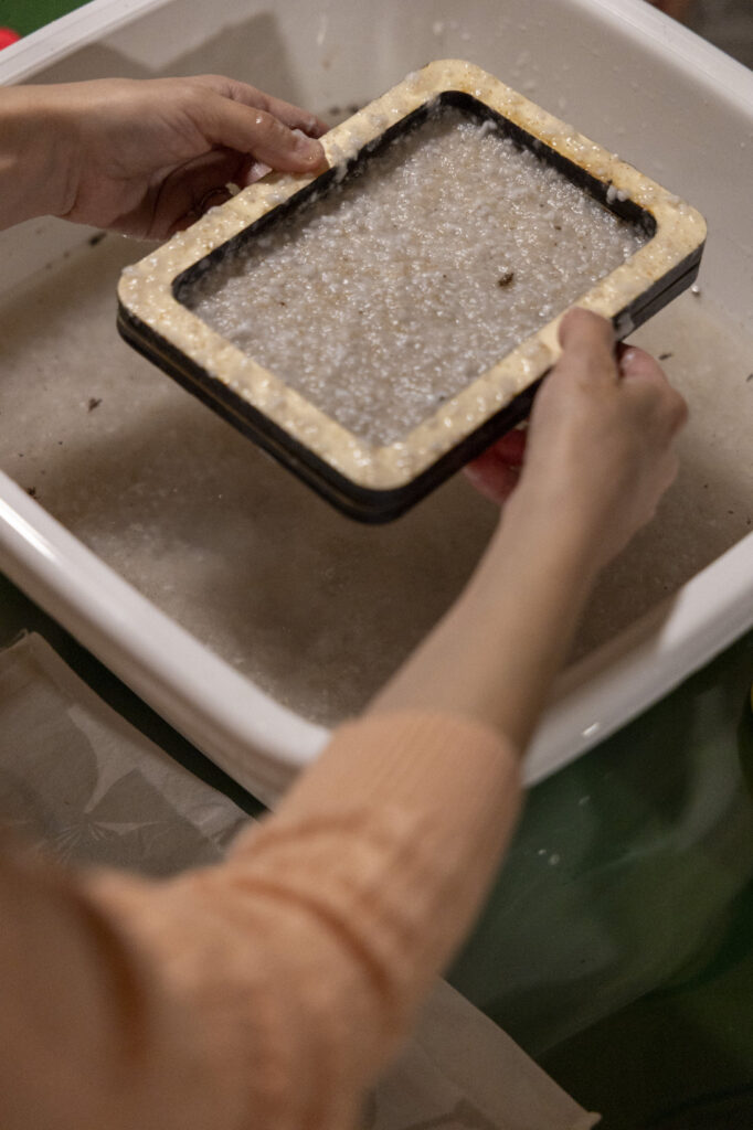 Formazione del foglio su telaio – laboratorio sCARTA Mani che reggono un telaio con carta artigianale bagnata
