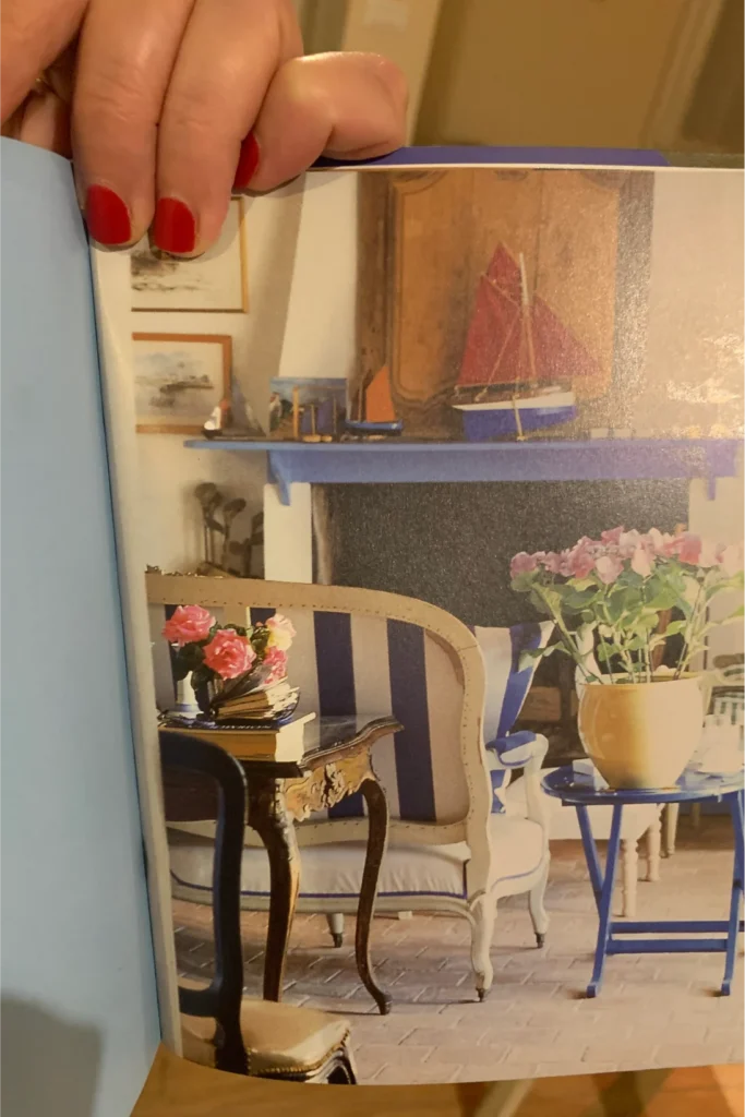 Soggiorno Mano con smalto rosso che tiene aperta una pagina di rivista raffigurante un interno con poltrona a righe blu e bianche, camino con modellino di barca a vela, vasi di fiori rosa e tavolino blu.