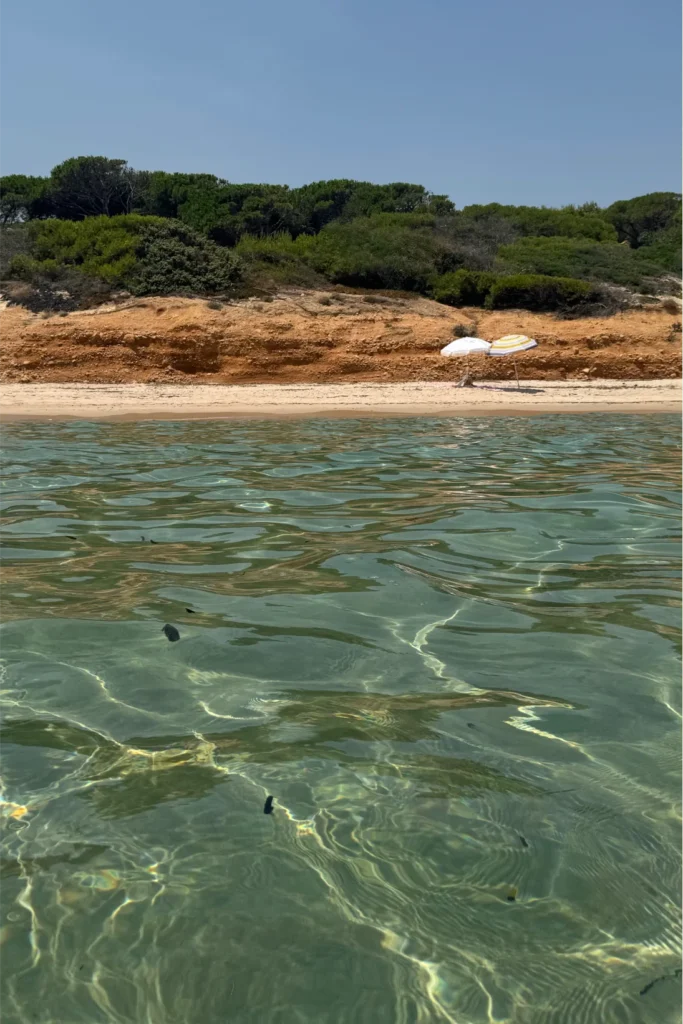 Spiaggia Vista dall'acqua verso una spiaggia sabbiosa con due ombrelloni, delimitata da una duna di terra rossastra e vegetazione mediterranea; acqua trasparente in primo piano.